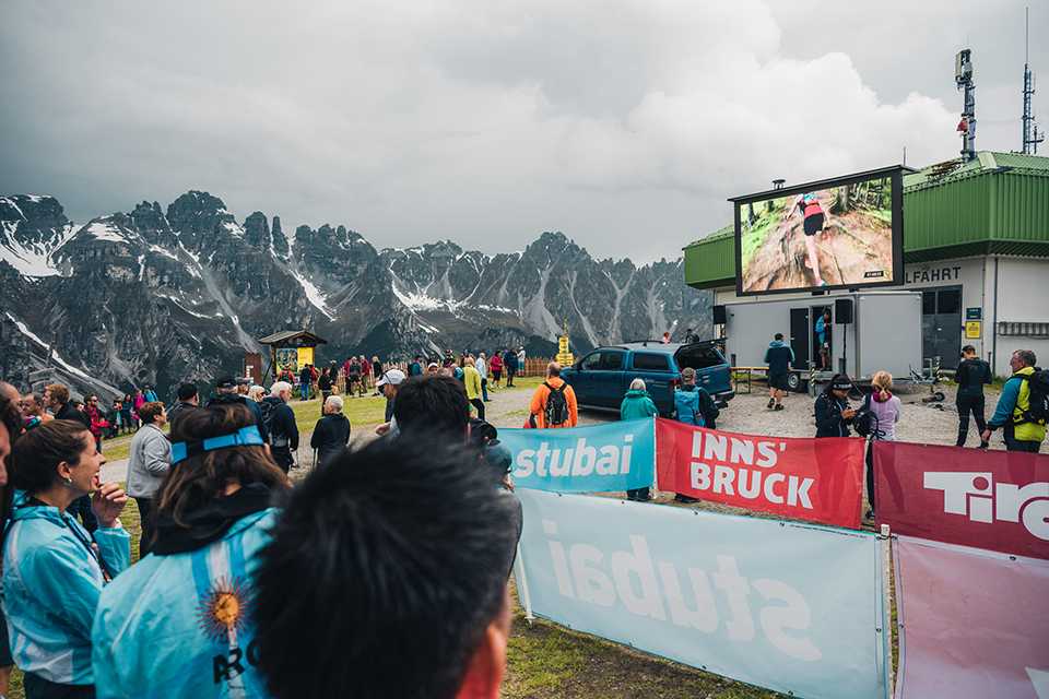 Trail Running WM Tirol 2023: Zuschauer verfolgen das Rennen auf einer Leinwand vor Bergkulisse.