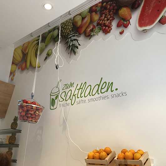 Saftladen Gebäude: Obst-Wandbild, Äpfel im Korb, Orangen in Kisten. Frische Säfte, Smoothies, Snacks.