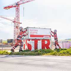 Baustelle Sonderbau: Bauarbeiter-Figuren mit Strabag-Werbung und Kran im Hintergrund.